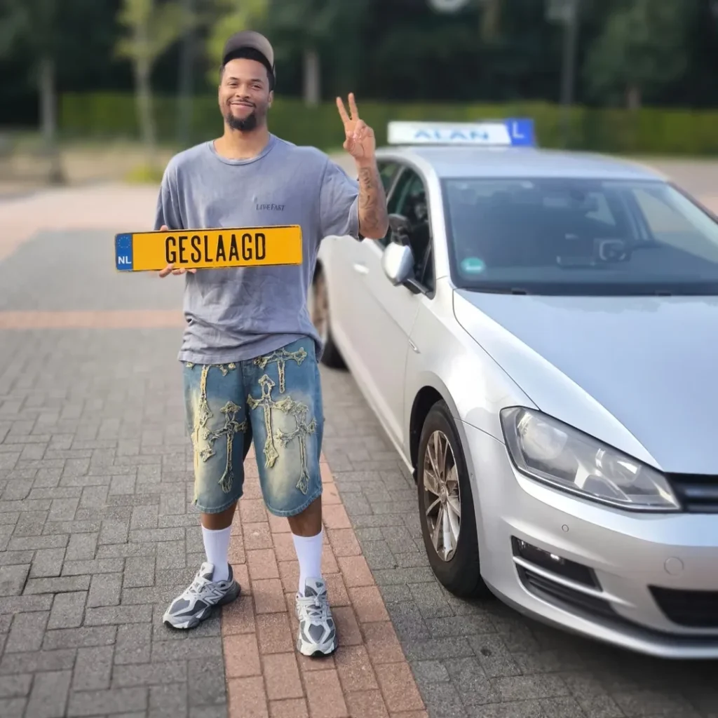 Andy geslaagd voor rijbewijs bij Autorijschool ALAN, staat met rijbewijsplaat naast witte auto en steekt overwinningsgebaar o Andy geslaagd voor rijbewijs bij Autorijschool ALAN, staat met rijbewijsplaat naast witte auto en steekt overwinningsgebaar o