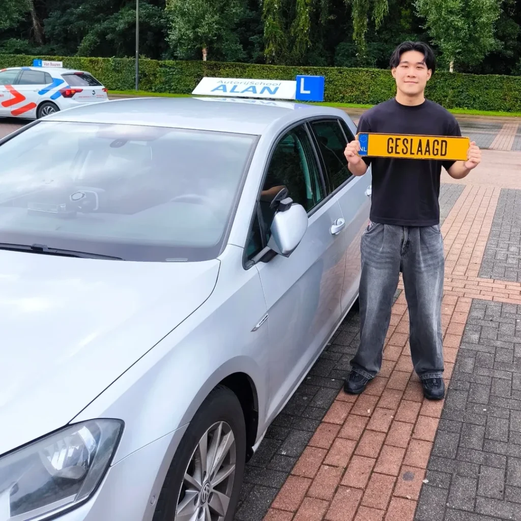 Benne houdt geslaagd kenteken vast naast witte auto bij Autorijschool ALAN na het behalen van zijn rijbewijs Benne houdt geslaagd kenteken vast naast witte auto bij Autorijschool ALAN na het behalen van zijn rijbewijs